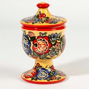 May include: A hand-painted, wooden jar with a lid. The jar is yellow with red trim and is decorated with red, blue, and white floral designs. The jar has a pedestal base and a rounded lid with a red knob.