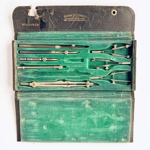 May include: A vintage Eugene Dietzgen Co. Reliance drafting instrument set housed in a black folding case with a green velvet interior. The set includes various metal compasses, dividers, and drawing tools secured in molded slots within the case.