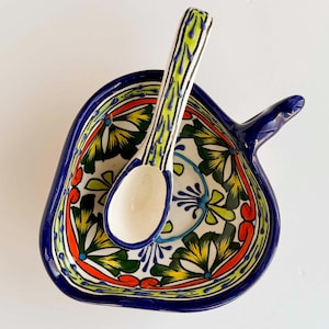 May include: A ceramic bowl shaped like a leaf with a matching spoon. The bowl features a hand-painted floral pattern in vibrant shades of blue, yellow, green, and orange against a white background, with a solid dark blue rim and handle.