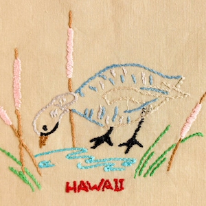 May include: A beige fabric panel featuring an embroidered scene of a bird standing in blue water near pink cattails and green grass. The bird is outlined in blue and white thread with black feet. The word HAWAII is embroidered in red below.