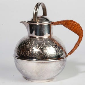 May include: A silver pitcher with a round body and a lid. The pitcher has an ornate floral pattern and a woven brown handle. The handle curves upward, and the lid has a small loop handle. The pitcher is on a white background.