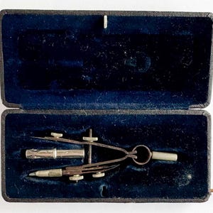 May include: A vintage drafting set in a black case with a blue velvet interior. The set includes a compass, a pen holder, and a small cylindrical piece. The case is rectangular with a hinged lid and a metal clasp.