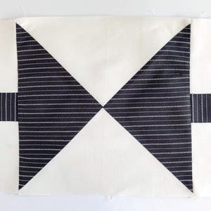 May include: A rectangular fabric quilt block featuring a central hourglass design composed of two black fabric triangles with thin white horizontal pinstripes, flanked by solid off-white fabric sections and horizontal black pinstriped strips.