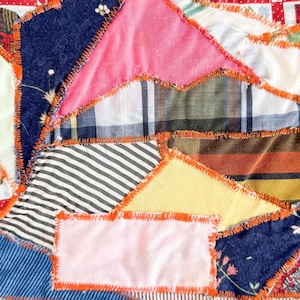 Op de afbeelding: Close-up van een patchworkquilt met verschillende stofpatronen en kleuren. De quilt heeft een mix van stoffen, waaronder strepen, ruiten en bloemenprints, aan elkaar gestikt met oranje draad. Het ontwerp is een mix van geometrische vormen.