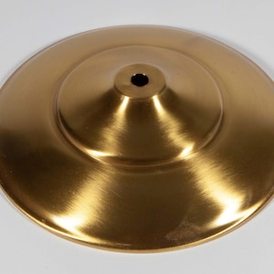 Puede incluir: Un objeto circular metálico de color dorado, con un cono central elevado y un pequeño agujero en la parte superior. La superficie tiene una textura cepillada, que refleja la luz. El objeto parece ser un elemento decorativo o parte de un accesorio más grande.