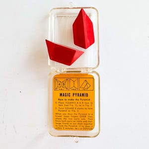 May include: A vintage "Magic Pyramid" puzzle game in a clear plastic case. The game includes two red triangular pieces and an instruction card with diagrams. The card has the text "MAGIC PYRAMID" and instructions on how to solve the puzzle.