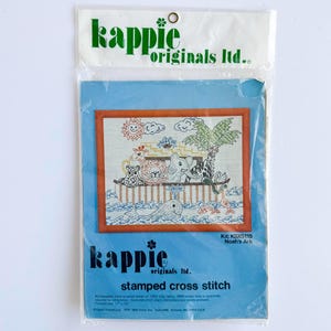 May include: A packaged cross-stitch kit featuring a colorful Noah's Ark design. The kit includes a stamped pattern with images of animals, the ark, and the sun. The packaging is blue and white with the brand name "kappie originals ltd." printed on it.