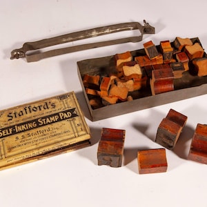 Op de afbeelding: Een vintage Stafford's stempelkussendoos, een metalen bakje met talloze kleine houten letterstempels en een losse metalen handgreep. De doos heeft zwarte tekst op een beige achtergrond, inclusief de merknaam en productdetails.