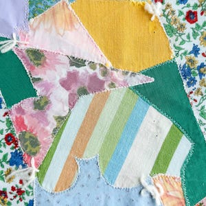 May include: A close-up of a patchwork textile with various fabric patterns. The design includes floral prints, stripes, and solid colors like green and yellow. White thread accents the seams, creating a handcrafted look.
