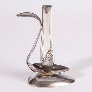 May include: A vintage silver bud vase with a unique design. The vase is crafted from repurposed silverware, featuring a slender, fluted body and floral detailing. A curved spoon-like element extends from the base, adding an artistic touch. The vase stands approximately 15 cm tall.