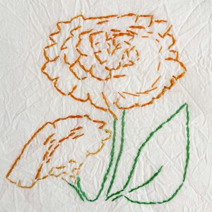 May include: Embroidered floral design on white fabric. The flower is orange with a green stem and leaves. The embroidery is done with a simple stitch, creating a textured effect.