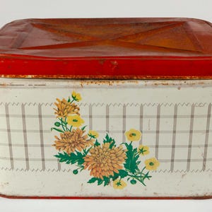 Puede incluir: Caja de lata rectangular vintage con tapa roja y cuerpo blanco. El cuerpo presenta un patrón de cuadros y un diseño floral con flores amarillas y naranjas. La caja tiene un aspecto rústico y envejecido.