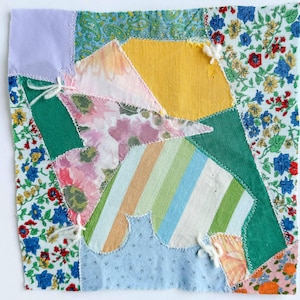 May include: A square fabric quilt block featuring a patchwork of various textile patterns including floral prints, solid yellow, light blue, and green sections, along with a striped panel, all joined together with white zigzag stitching.