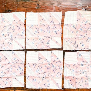 May include: Six square fabric quilt blocks arranged in two rows of three. Each block features a geometric star pattern constructed from alternating panels of light pink fabric with a white floral print and off-white fabric with thin stripes.
