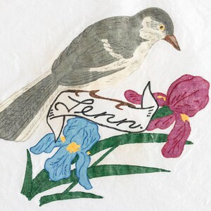 May include: A watercolor illustration on white paper featuring a grey and white bird perched above a banner with the handwritten text "Lenn." Below the banner are two iris flowers, one blue and one magenta, with green leaves and stems.