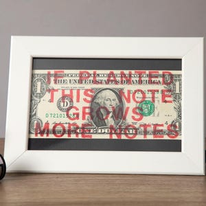 May include: A framed one-dollar bill with the text "If planted, this note grows more notes" in red. The white frame sits on a wooden surface, with a film roll and green sunglasses nearby.