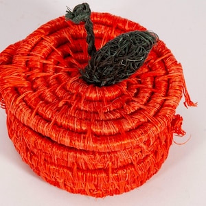 May include: A woven red apple-shaped basket with a dark green stem and leaf. The basket is made of a tightly woven material, and the lid fits snugly on the base. The apple basket is a decorative storage item.