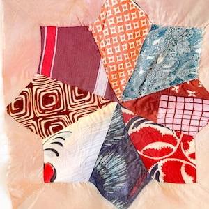 May include: A square fabric quilt block featuring a central star design composed of various patterned textile patches in shades of orange, blue, red, white, and dark gray, all framed by a solid light peach-colored fabric border.