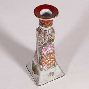 May include: A decorative ceramic candlestick holder with a red rim and floral design. The candlestick has a square base and a flared body, decorated with pink and orange flowers and green leaves.