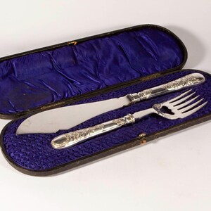 Könnte beinhalten: Ein silbernes Messer- und Gabel-Set mit floralen Details, präsentiert in einem offenen Vintage-Etui mit tiefblauem Samtfutter. Das Messer hat eine breite Klinge, und die Gabel hat vier Zinken.