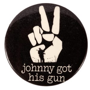May include: A black round button with a white peace sign and the text "johnny got his gun". The button features a silhouette of a person in the peace sign.
