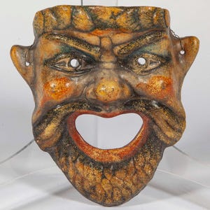 May include: A detailed, antique-style mask with a stern expression. The mask features a prominent nose, a wide open mouth, and detailed facial features. The color palette includes shades of brown, orange, and gold, with accents of green and red.