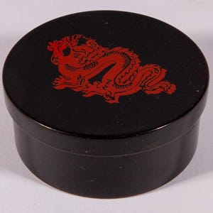 May include: A black, round box with a red dragon design on the lid. The dragon is stylized and detailed, set against the glossy black surface. The box is cylindrical with a matching black base.
