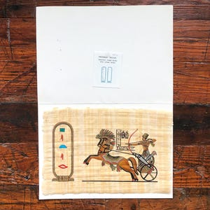 Puede incluir: Pintura egipcia sobre papiro que representa una escena de carro con un caballo, un arquero y jeroglíficos. La obra de arte utiliza tonos cálidos de marrón, rojo y dorado. La pintura está montada sobre un fondo blanco, con una pequeña etiqueta impresa.