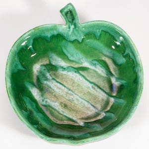 Könnte beinhalten: Eine grüne, keramikförmige Apfelschale mit glänzendem Finish. Die Schale hat einen Stiel und ein strukturiertes Inneres mit Grüntönen, Weiß und Beige. Das Design erinnert an einen echten Apfel.