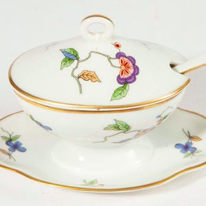 Può includere: Un piatto da portata in porcellana bianca con coperchio, cucchiaio e piattino. Il piatto presenta disegni floreali in viola, arancione e blu, con finiture dorate. Il set è progettato per servire salse o condimenti.