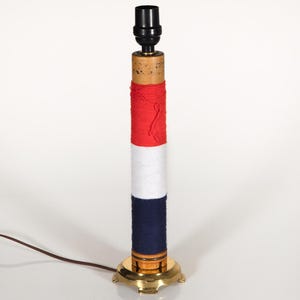 May include: A handmade table lamp with a cylindrical base wrapped in red, white, and navy blue yarn. The lamp has a brass base and a black lamp holder. The cord is brown.
