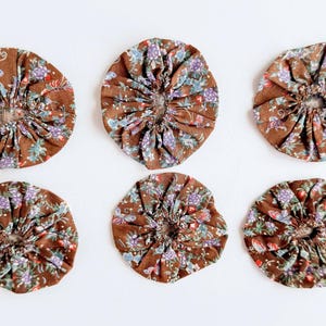 May include: Six circular fabric yo-yos arranged in two rows. Each piece is made from brown cotton fabric featuring a repeating floral pattern with small purple, red, and green flowers. The fabric is gathered in the center to create a ruffled effect.