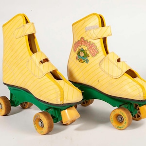 Könnte beinhalten: Paar gelbe Vintage-Rollschuhe mit grünem Fahrgestell und orangefarbenen Rollen. Die Rollschuhe haben ein vertikales Streifenmuster und das Logo "Cabbage Patch Kids". Die Räder haben einen Durchmesser von ca. 5 cm.
