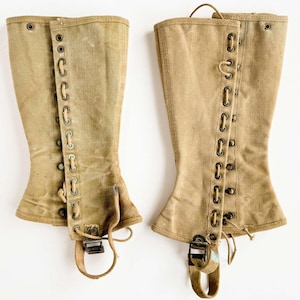 May include: A pair of vintage tan canvas gaiters. They have metal eyelets for lacing, buckle closures, and straps. These gaiters are designed to protect the lower legs and are a classic piece of military or outdoor gear.