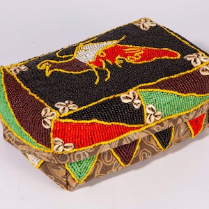 May include: A rectangular box featuring a lid covered in black, red, green, and yellow glass beads with a central bird motif. The lid is accented with small white cowrie shells. The base is wrapped in brown fabric with a tan swirling pattern.