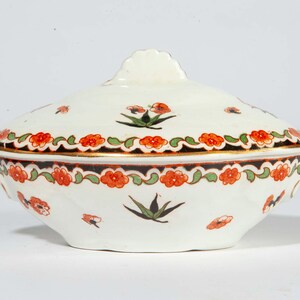 May include: A white ceramic serving dish with a lid, featuring a floral design in orange, green, and black. The dish has a gold trim and small handles on either side. The lid has a decorative finial.
