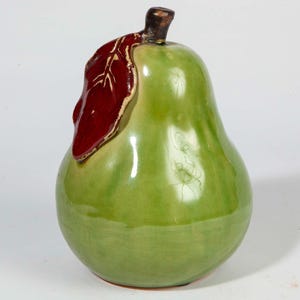 May include: A ceramic pear figurine in shades of green with a dark red leaf and a brown stem. The pear has a glossy finish and is a decorative item.