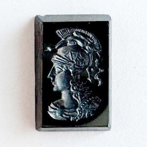 May include: A rectangular, black cameo featuring a profile of a figure wearing a helmet and armor. The figure is carved in a light gray color, set against a dark background. The cameo is framed with a dark gray border.