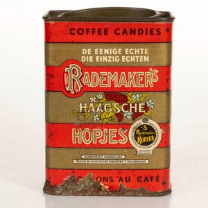 May include: Vintage Rademaker's Haagsche Hopjes coffee candy tin. The rectangular tin features a red and gold color scheme with text in multiple languages. The tin has a hinged lid and shows signs of age and wear.