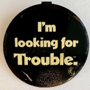 Könnte beinhalten: Ein runder, schwarzer Knopf mit dem Text "I'm looking for Trouble" in einer fetten, cremefarbenen Schrift. Der Knopf hat eine kleine, rechteckige Befestigung oben und ein leicht abgenutztes Aussehen.