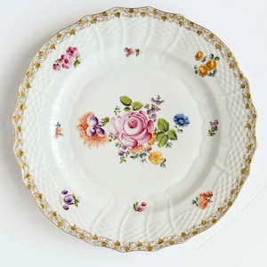 Puede incluir: Plato de porcelana blanca con diseño floral. El plato presenta una disposición central de flores rosas, moradas y amarillas, rodeadas de grupos florales más pequeños. Un borde dorado y ornamentado añade un toque decorativo.
