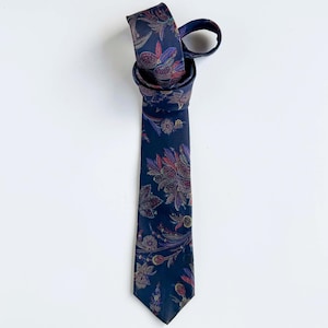 May include: A dark navy blue necktie featuring a repeating floral and paisley pattern in shades of muted red, purple, gold, and green. The tie is knotted at the top, with the long blade hanging straight down, displaying the intricate botanical print.