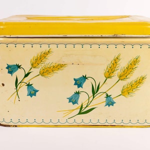 Op de afbeelding: Een rechthoekige vintage blikken doos met een gele deksel en een crèmekleurige body. De doos is versierd met illustraties van tarwe en blauwe klokjesbloemen. De randen zijn afgewerkt met een geschulpte rand.