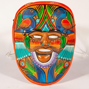 May include: A hand-painted decorative mask featuring a central human face with a mustache and open mouth. The design includes two blue birds at the top, vibrant green, red, and turquoise geometric patterns, and a bright orange border.