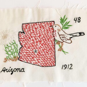 May include: A rectangular piece of off-white fabric featuring an embroidered red map of Arizona with a small star, a white cactus flower, a brown and white bird perched on green branches, and the black embroidered text Arizona, 48, and 1912.