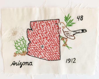 May include: A rectangular piece of off-white fabric featuring an embroidered red map of Arizona with a small star, a white cactus flower, a brown and white bird perched on green branches, and the black embroidered text Arizona, 48, and 1912.