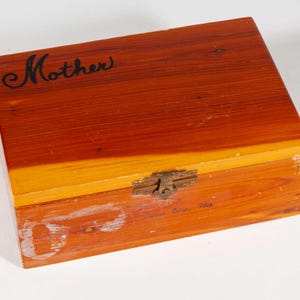 Può includere: Una scatola rettangolare di legno con coperchio incernierato. La scatola è tinta in tonalità arancioni e marroni. La parola "Mother" è scritta in corsivo sulla parte superiore. Un fermaglio in metallo fissa il coperchio.