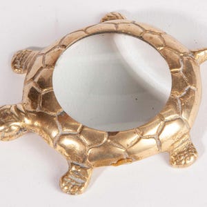 May include: A gold-colored turtle-shaped magnifying glass. The magnifying glass has a clear, circular lens in the center, surrounded by a detailed turtle design. The turtle's shell and limbs are textured, and the overall design is ornate.