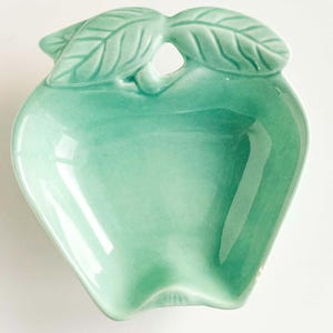 May include: A light mint green ceramic dish shaped like an apple. The bowl features a smooth, glossy finish with a molded leaf detail at the top center. The interior is concave, and the edges are curved to mimic the silhouette of a piece of fruit.