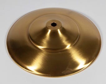 May include: A circular, brushed gold metal base with a smooth, slightly domed profile and a centered, circular opening. The metallic surface features a subtle, directional grain texture that reflects light across its curved, wide, flat rim.